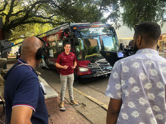 Image of Blake in front of a USC Transit bus where he is speaking to a camera. Producers can be seen holding a microphone above him,